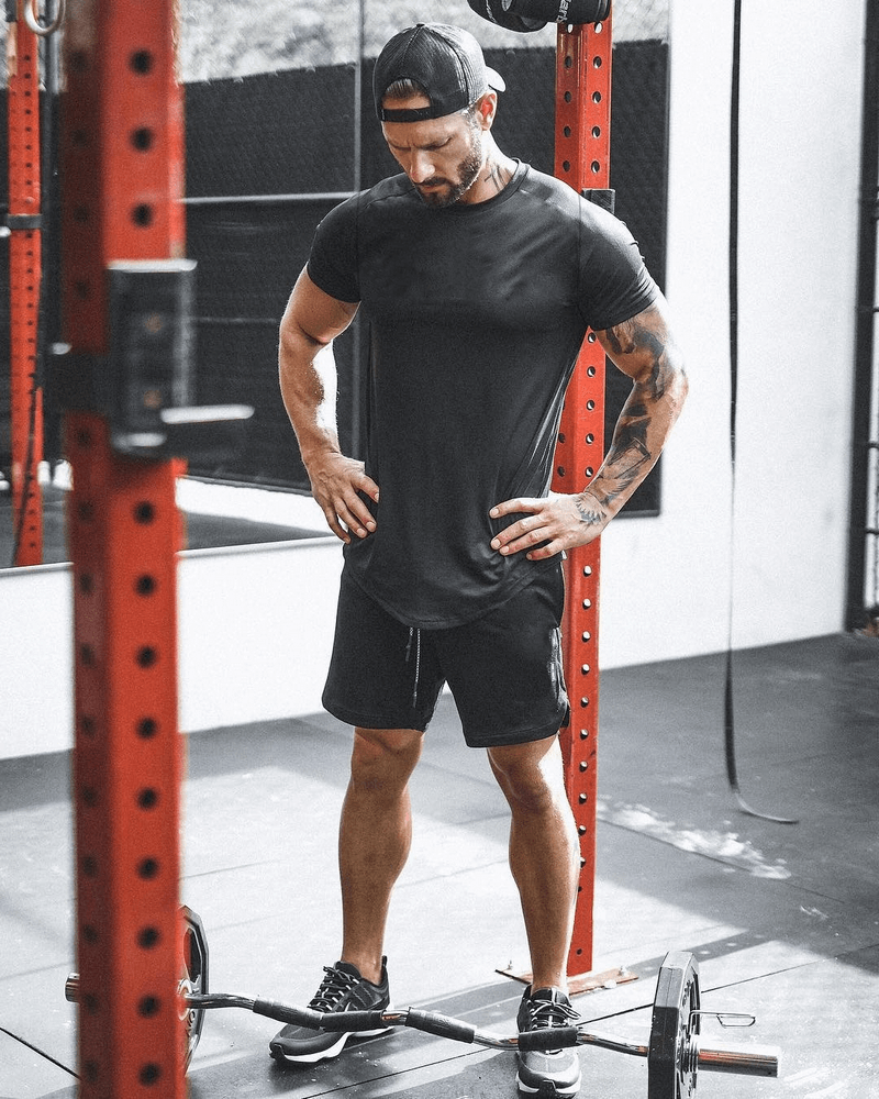 Man wearing a black o-neck short-sleeve jersey t-shirt, standing in a gym with fitness equipment around him.